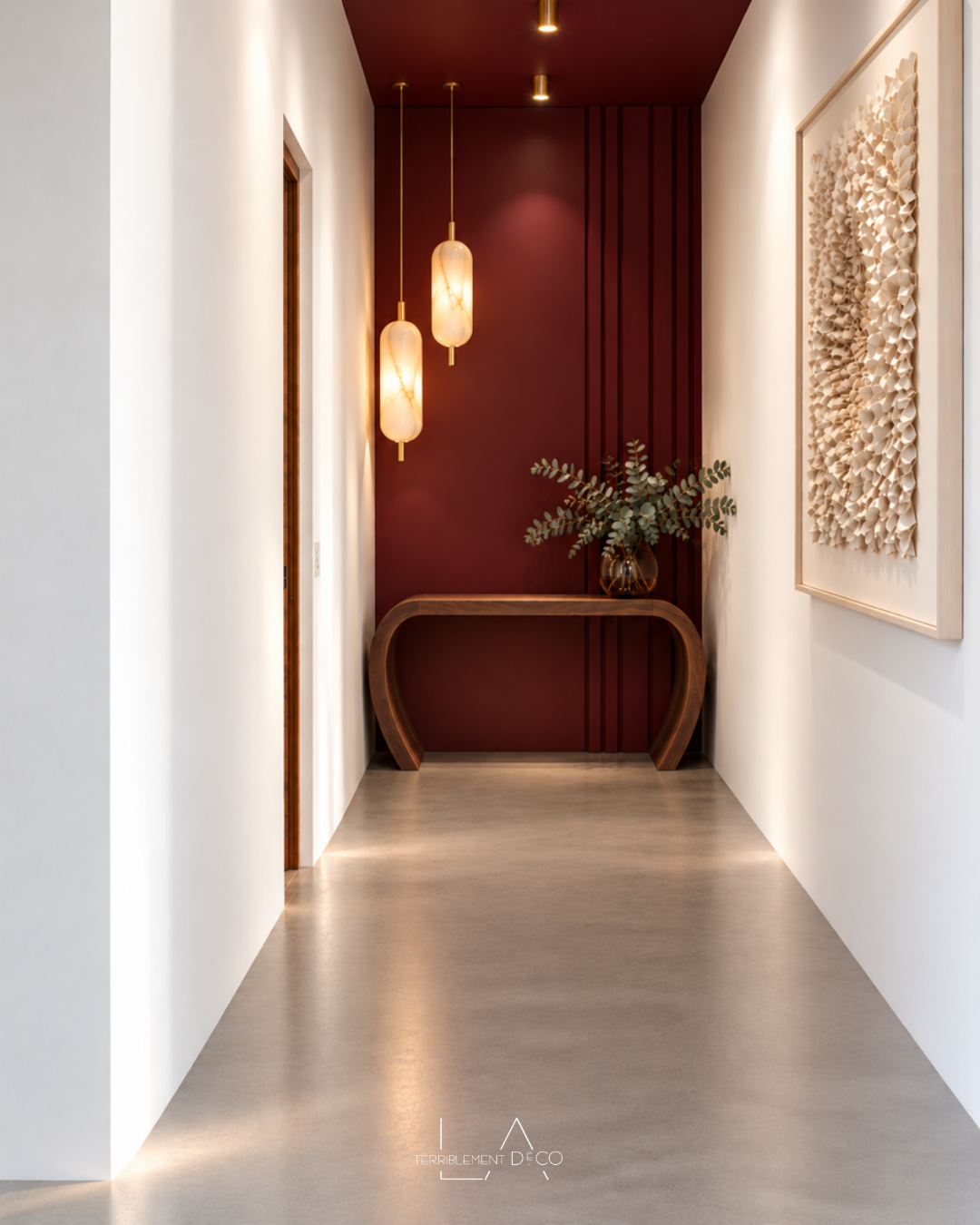 Fond du couloir : aménagement avec un mur rouge bordeaux, une console sculpturale en bois, deux suspensions lumineuses et un tableau contemporain sur le mur latéral, un design de Laurence ARNAUD